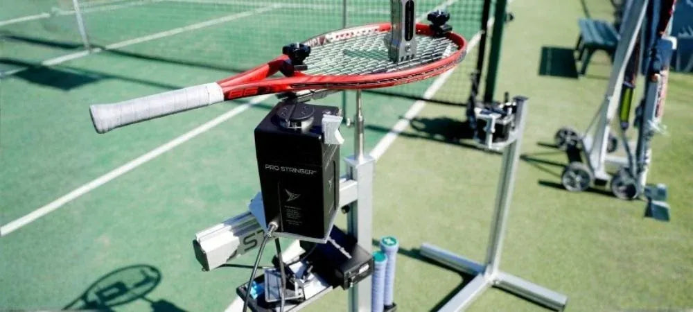 Een racket met rood frame gemonteerd op een draagbare bespanmachine op een standaard naast een tennisbaan