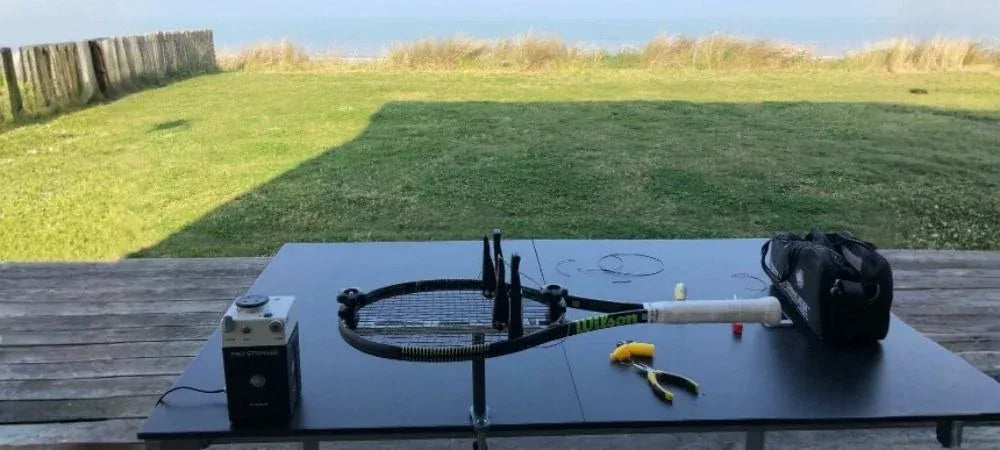 Een racket gemonteerd op een draagbare bespanmachine op een tafel buiten, met een tang en een tas ernaast